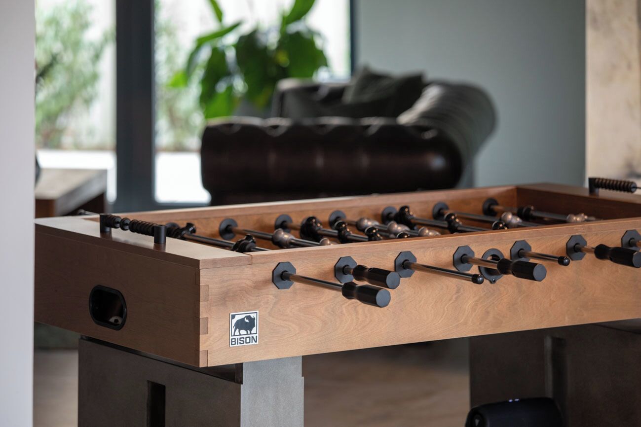 Une table de baby-foot en bois ornée du logo Bison, placée dans une pièce où se trouvent un canapé en cuir et une plante en arrière-plan.