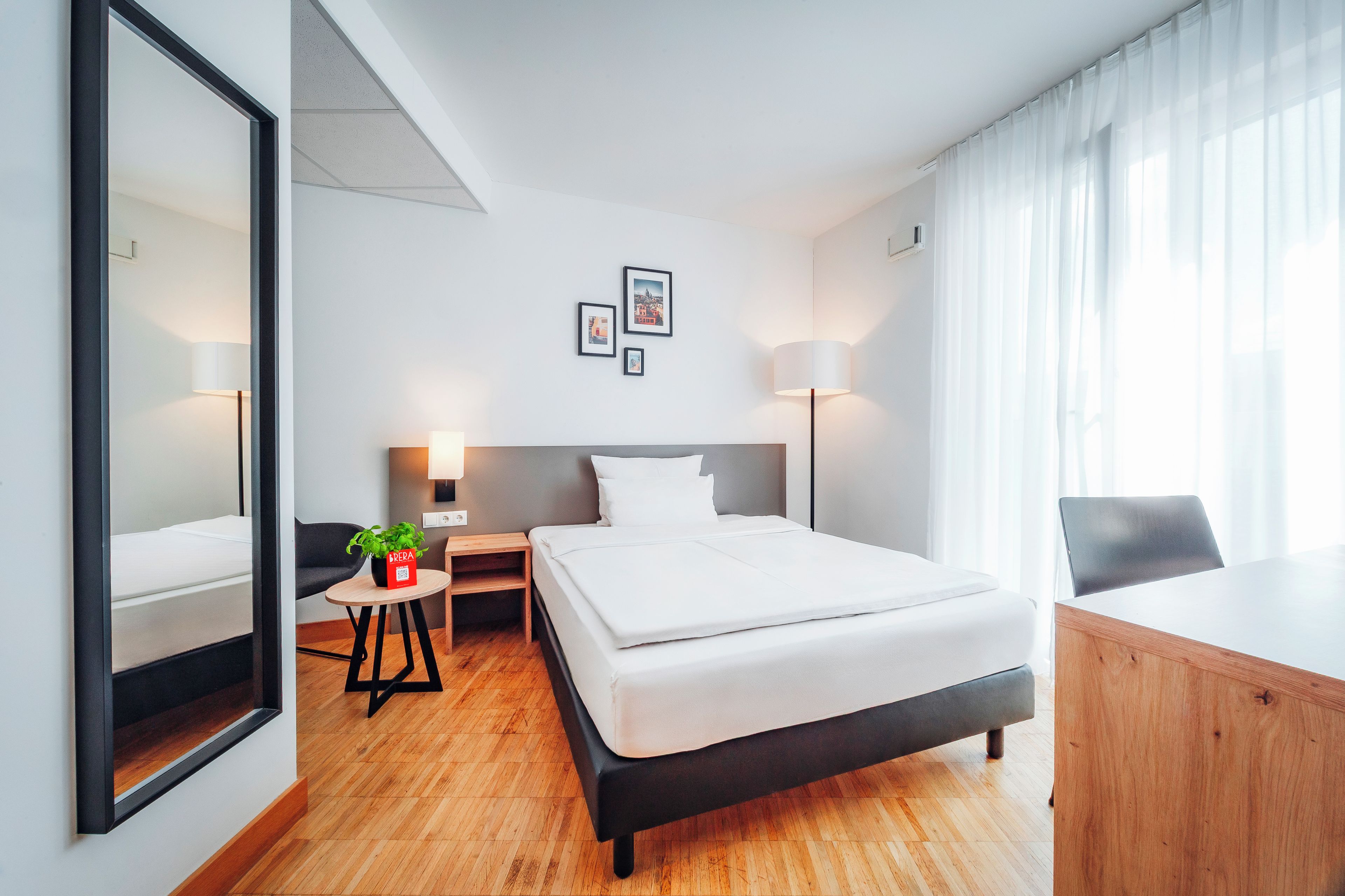 A modern hotel room with a large bed, a desk, a chair, a small table with a plant, and a large mirror.