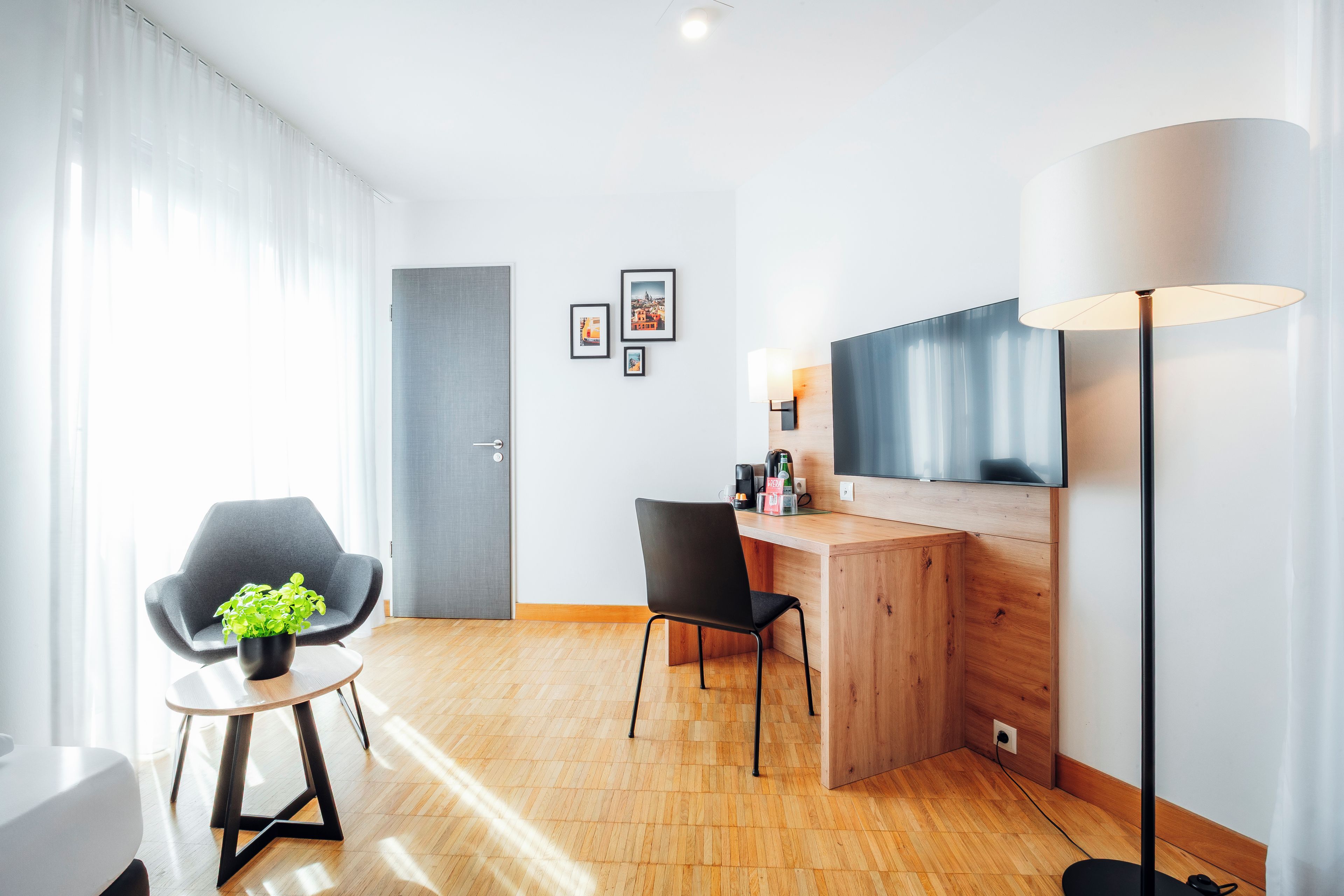 A modern hotel room with a wooden desk, a black chair, a small table with a plant, a large floor lamp, and a flat-screen TV mounted on the wall.