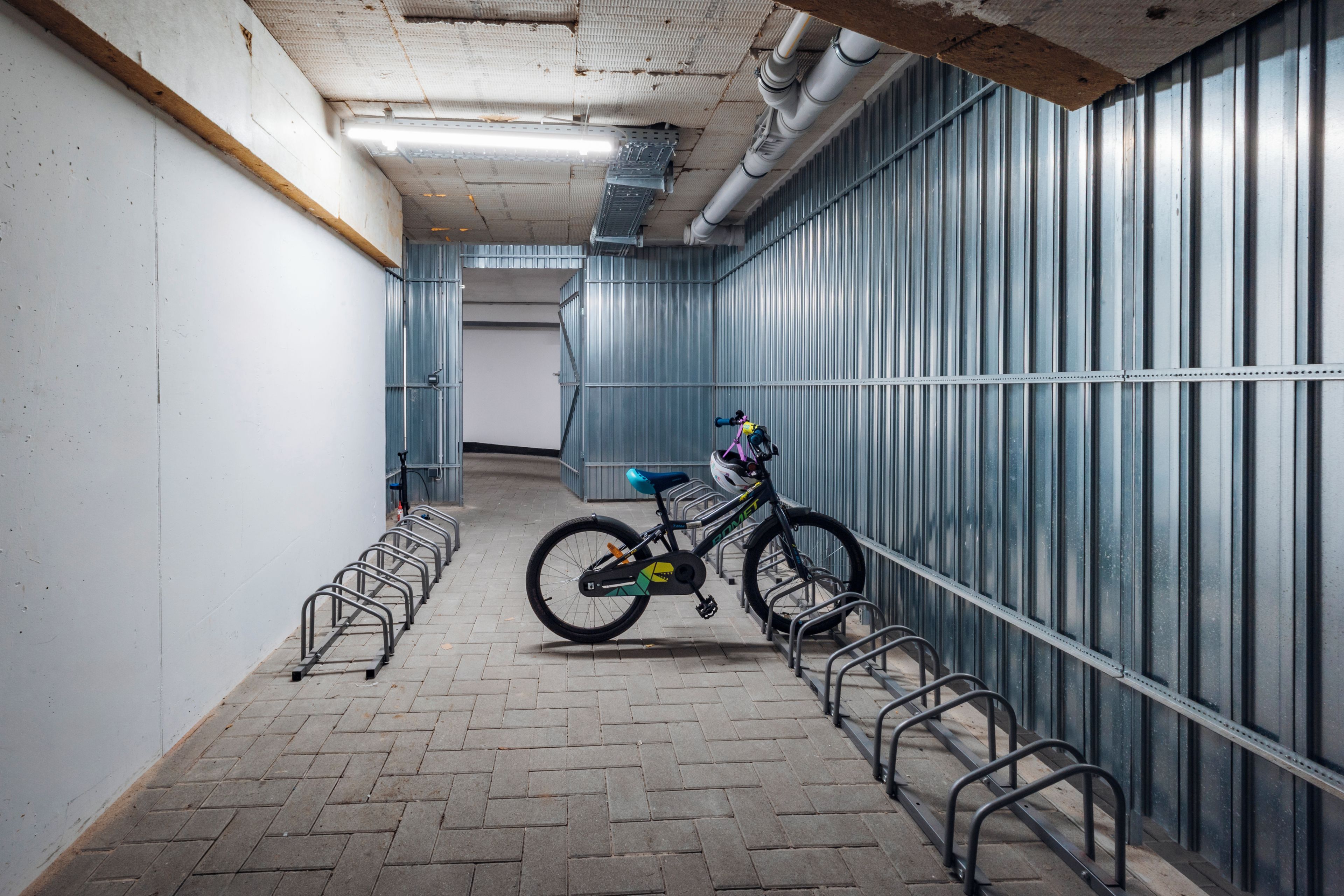 A bicycle parked in a metal rack, a tiled floor, and a corridor with metal walls and a ceiling with visible pipes.