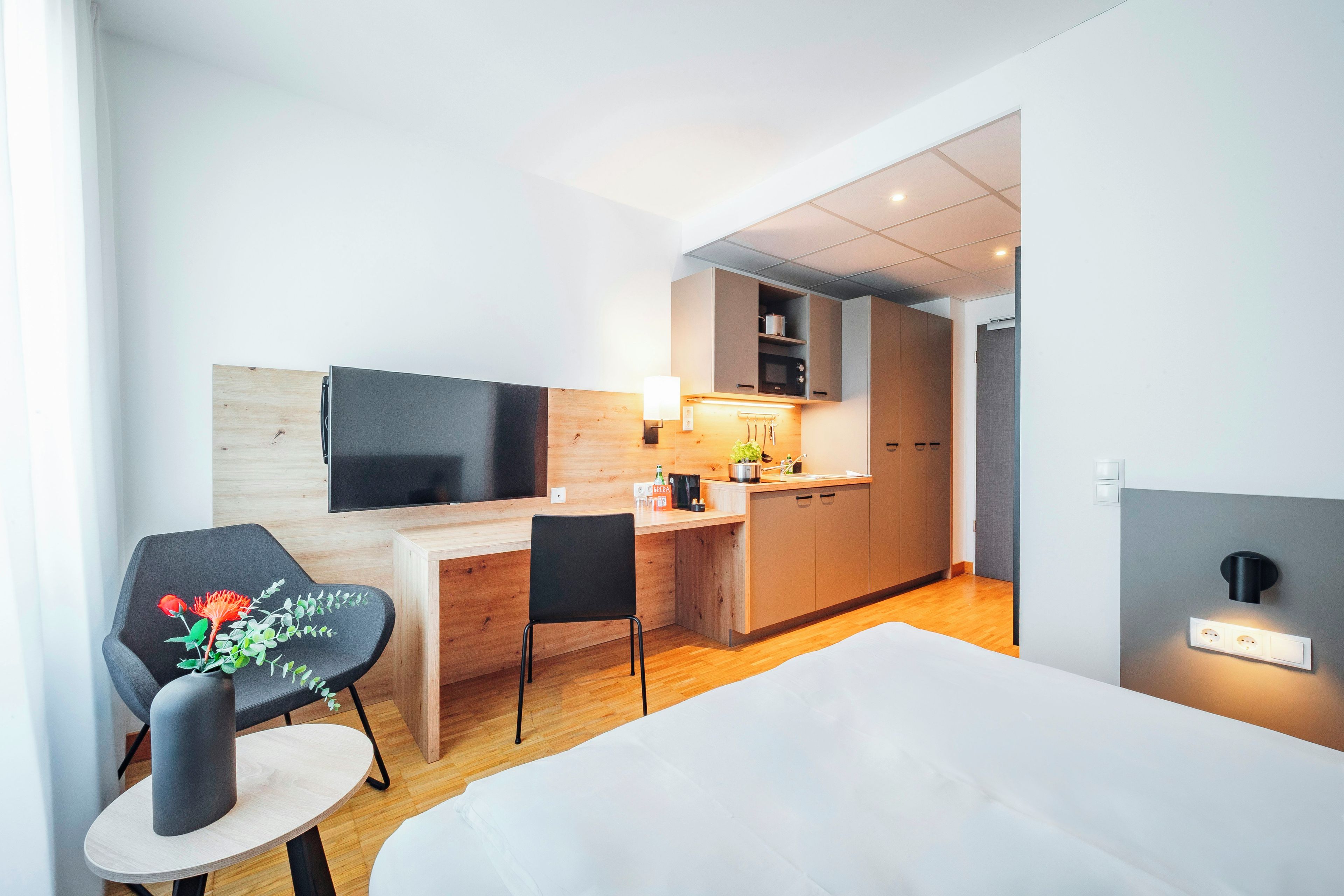 A modern hotel room with a bed, a desk, a chair, a television, and a kitchenette.