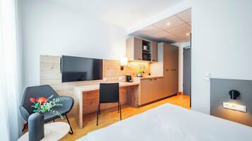 A modern hotel room with a bed, a desk, a chair, a television, and a kitchenette.