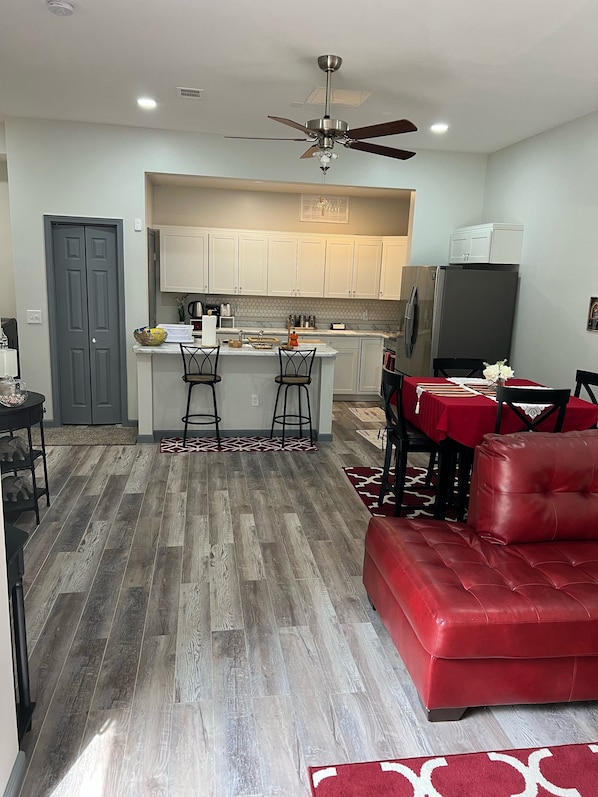 Una cocina moderna con armarios blancos, una isla central con taburetes, una zona de comedor con un sofá rojo y una puerta gris.