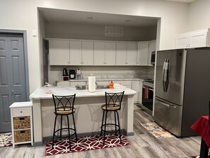 Una cocina moderna con armarios blancos, encimera de mármol, dos taburetes de bar, un refrigerador de acero inoxidable y una alfombra roja estampada.