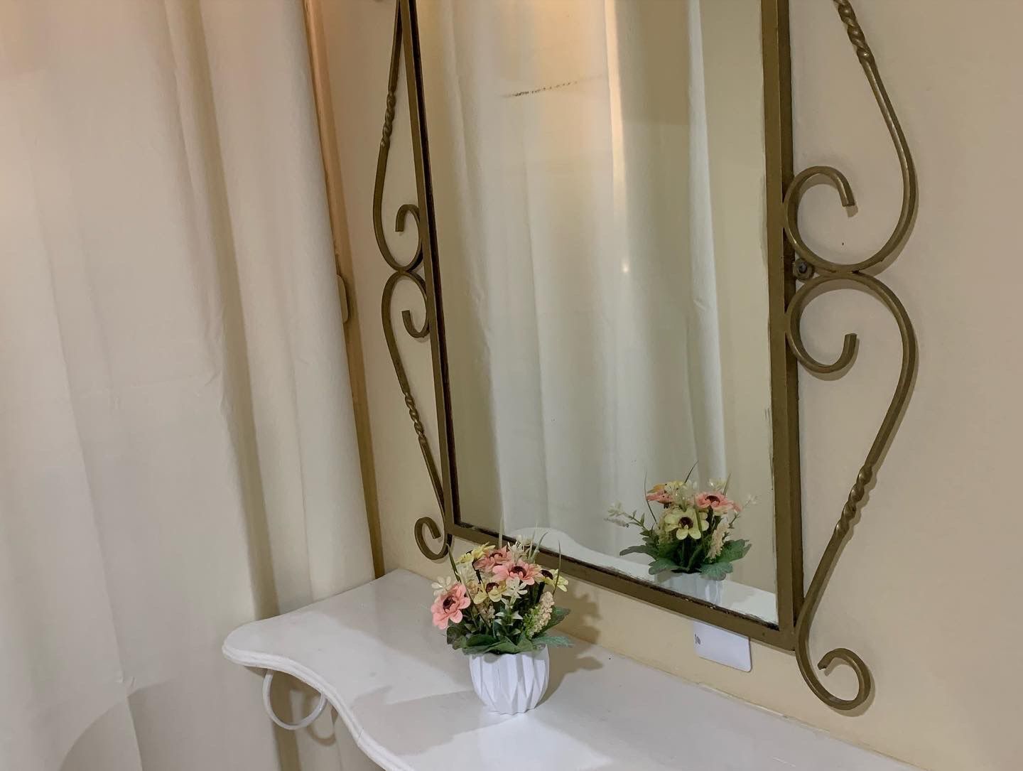 A bathroom vanity with a white countertop and a mirror with ornate gold framing. A small white vase with pink and yellow flowers is placed on the vanity.