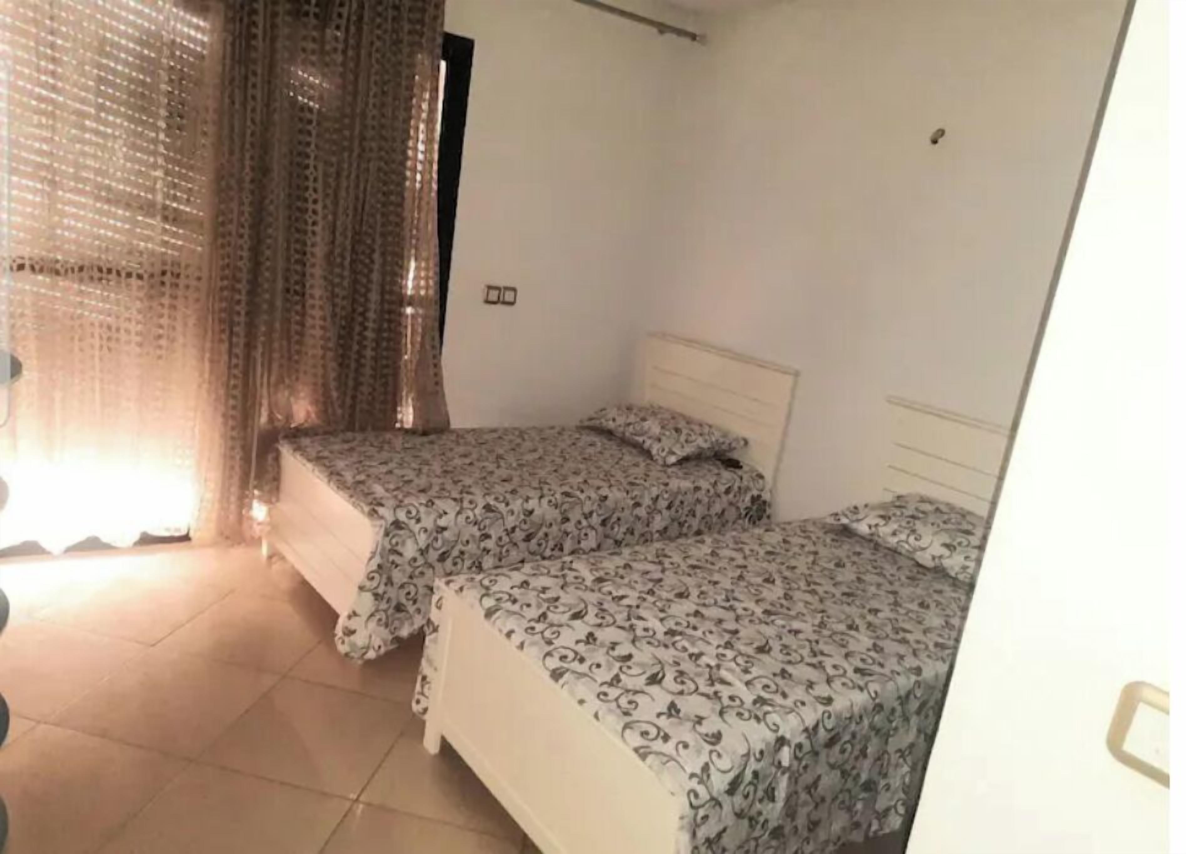 A room with two beds, a window with curtains, and a tiled floor.