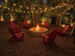 A fire pit surrounded by red chairs in a wooded area with string lights.