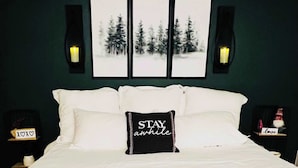 A bed with white bedding, a black throw blanket, and a pillow with the text "STAY awhile". There are two wall-mounted lamps and three framed pictures above the bed. - Spacious 4BR Near Pinehurst | Fire Pit + Games (Southern Pines)