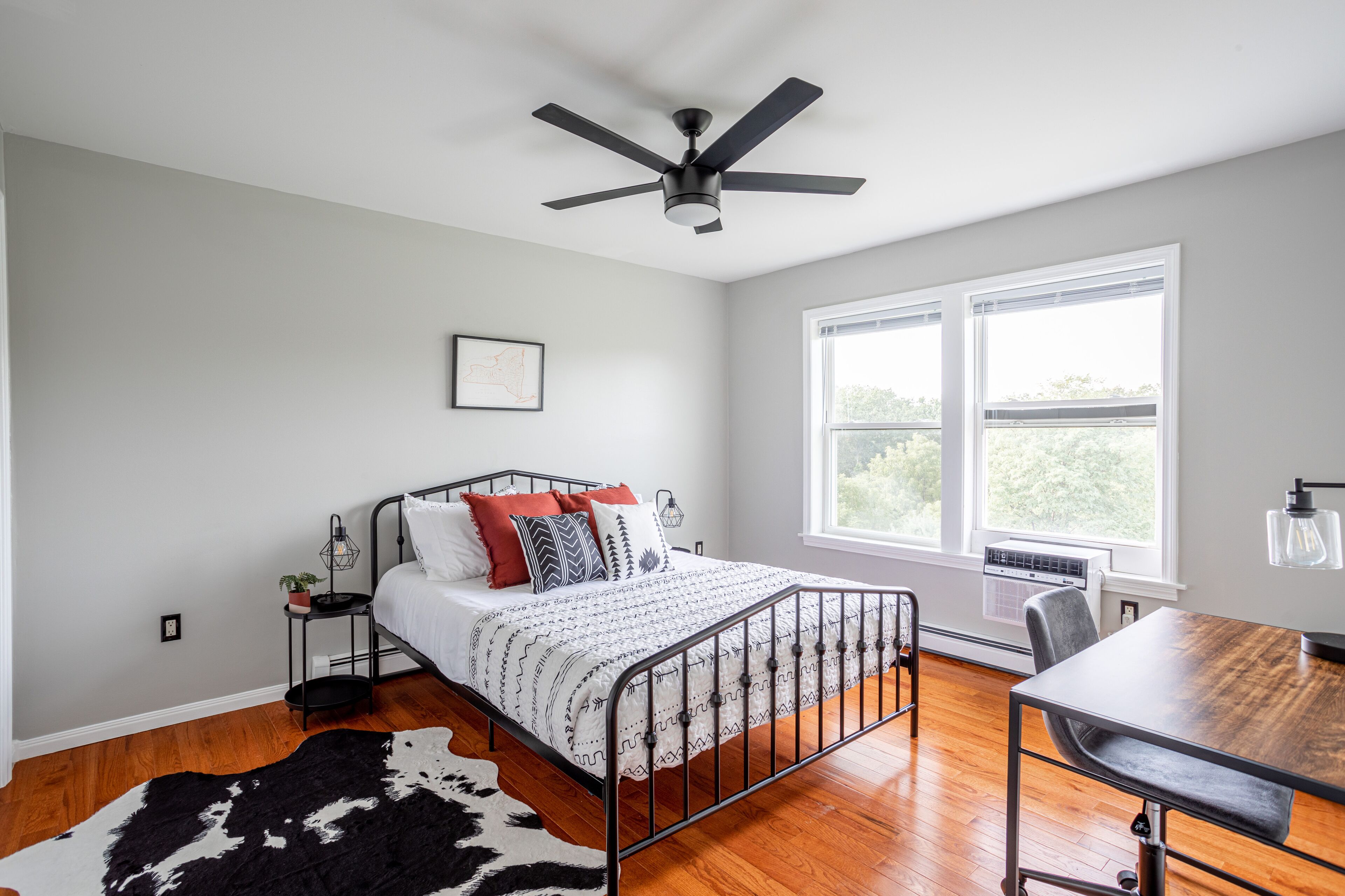 A bedroom with a bed, a desk, a chair, a ceiling fan, a window, and a cowhide rug.