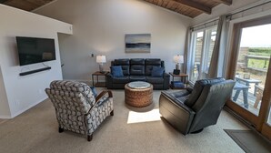 A living room with a large TV, a dark leather sofa, a patterned armchair, and a wicker ottoman.