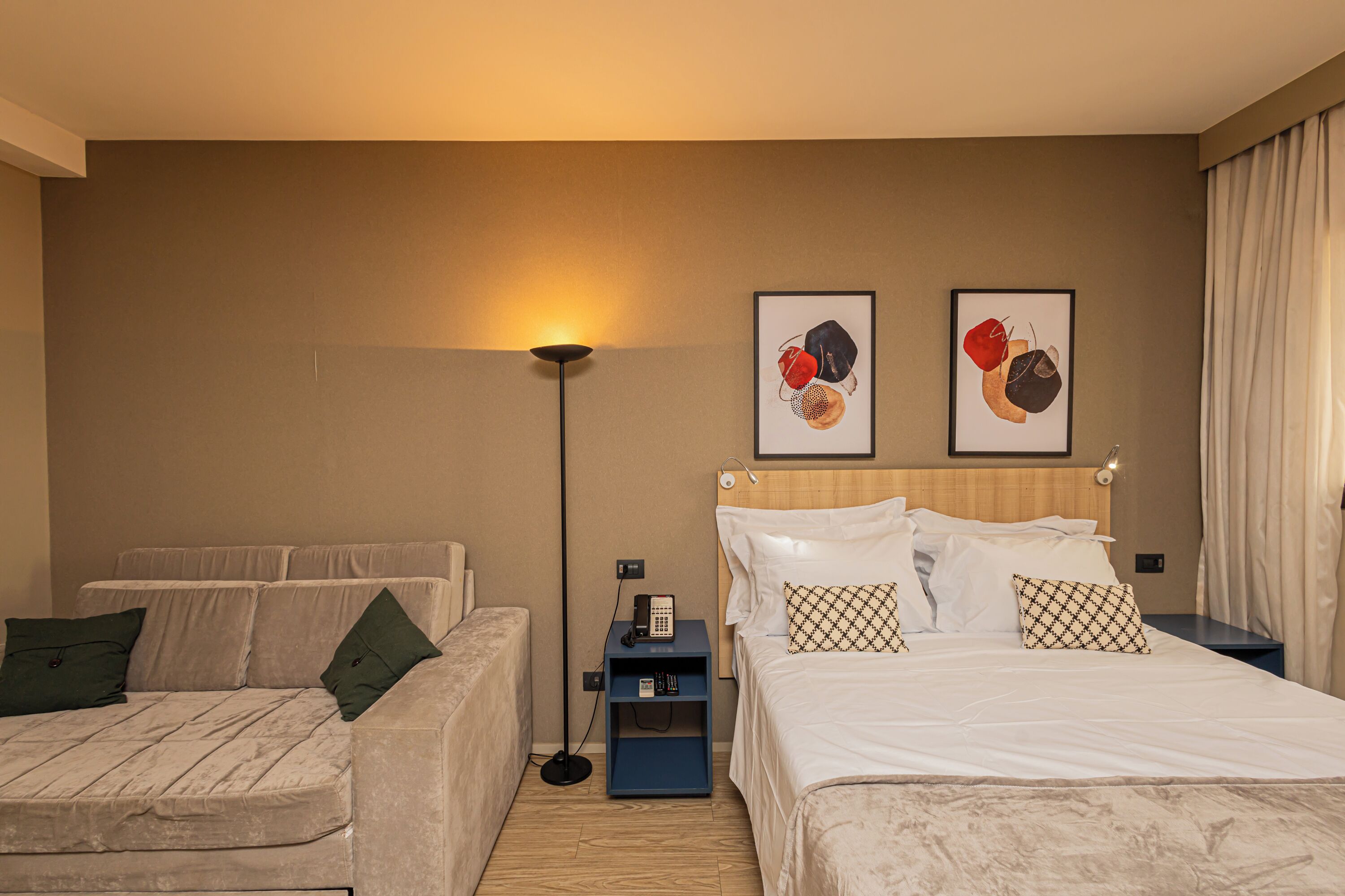 A hotel room with a bed, a sofa, a nightstand with a phone, and artwork on the wall.