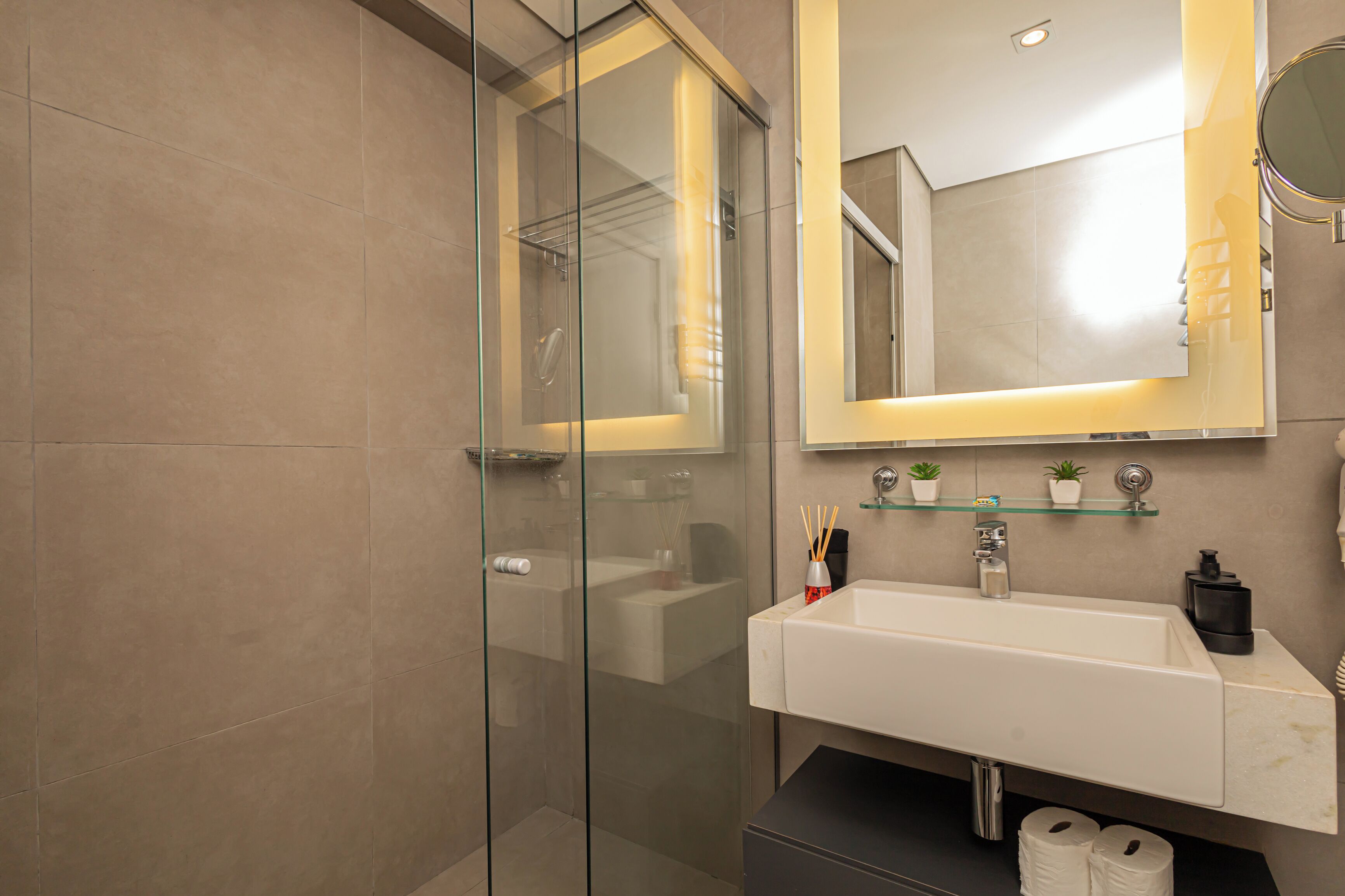 Un baño moderno con cabina de ducha de vidrio, un lavabo blanco e iluminación integrada en el espejo.