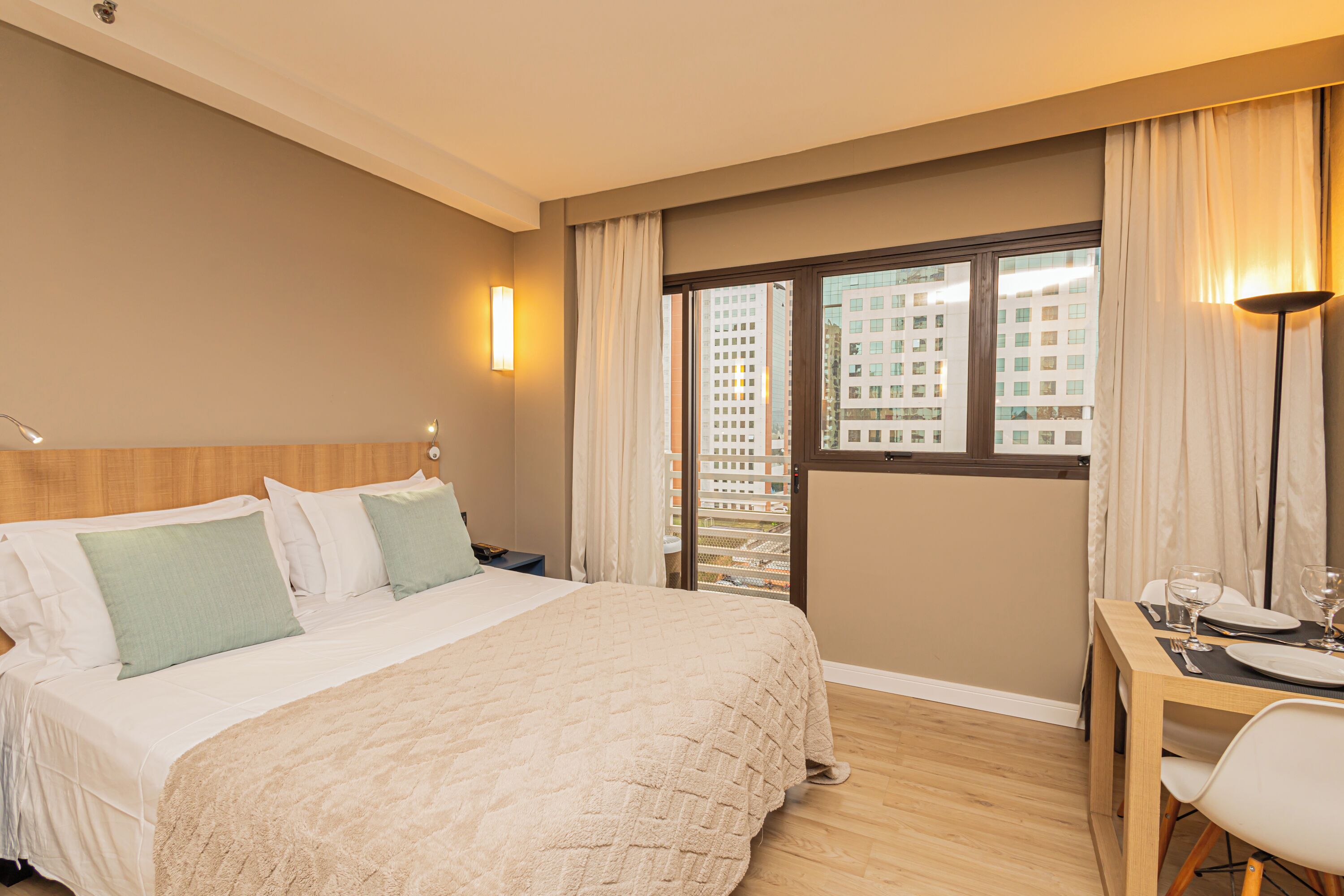 A hotel room with a bed, a dining table with chairs, a view of buildings through a window, and a lamp.