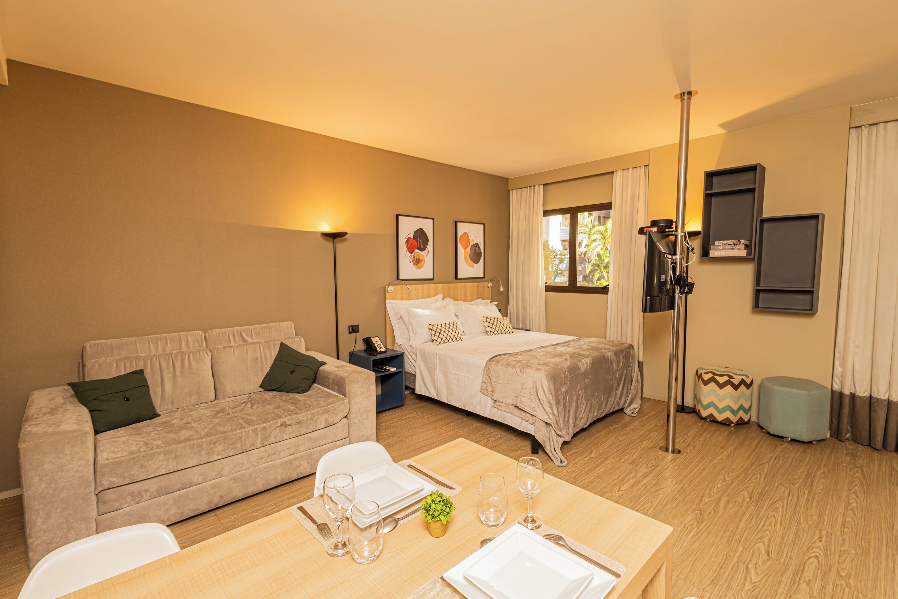 A hotel room with a bed, a sofa, a dining table set with plates and glasses, a lamp, and a window with curtains.