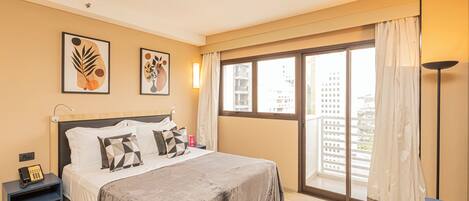 A hotel room with a bed, a nightstand, a desk, and a large window with a view of buildings.