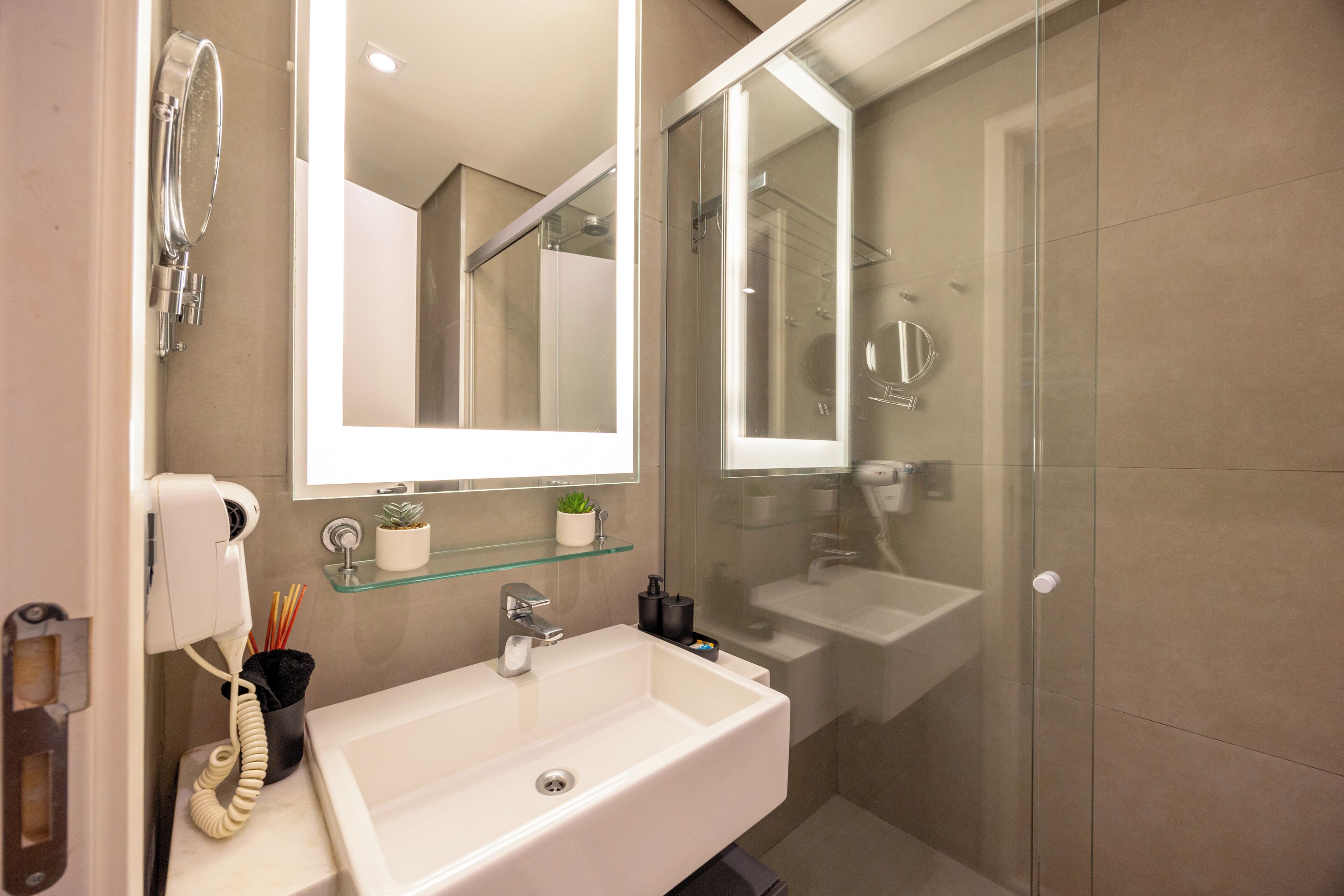 Un baño con lavamanos blanco, repisa de vidrio con una planta, espejo y cabina de ducha de vidrio.