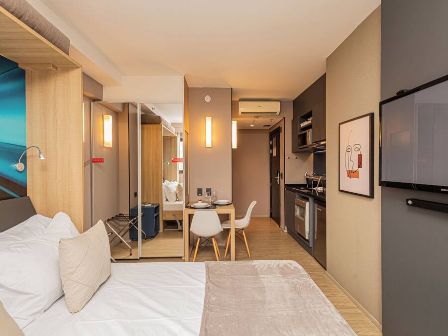 A hotel room with a bed, a desk, a chair, a TV, and a kitchenette.