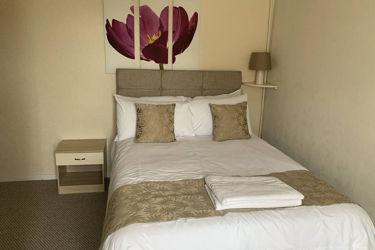 A neatly made bed with a white and beige floral bedspread, two beige pillows, and a folded white towel. A small bedside table with a drawer is visible to the left.