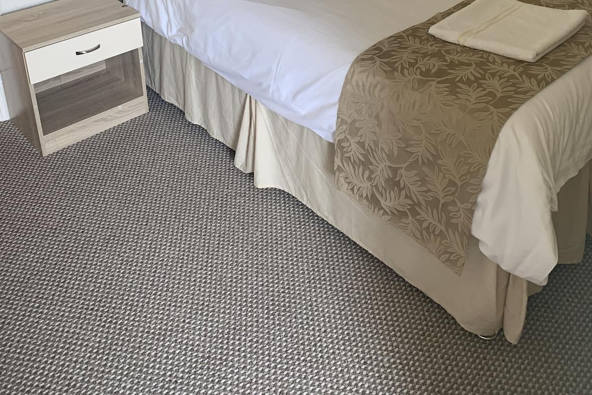 A neatly made bed with a headboard, a bedside table with a drawer, and a patterned carpet.