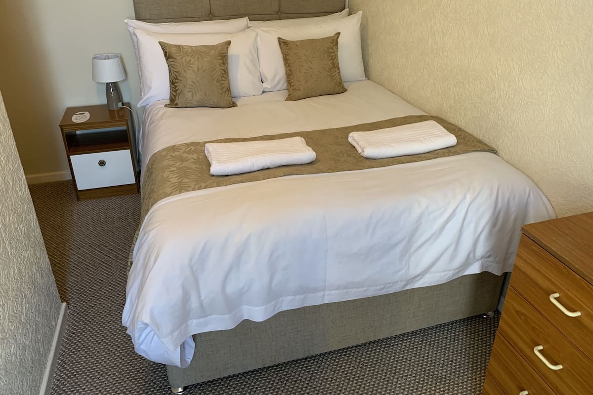 A neatly made bed with white and brown linens, a bedside table with a lamp, and a wooden nightstand.