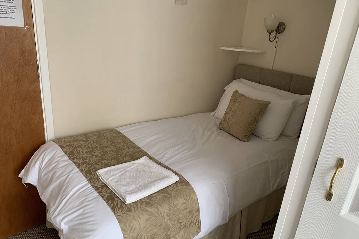 A single bed with a beige and white bedspread, a white pillow, and a beige pillow with a leaf pattern. There is a white towel on the bed.