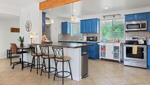 Une cuisine avec des meubles de couleur bleue, un îlot central doté de tabourets, un four à micro-ondes, un four traditionnel et une fenêtre avec des stores.