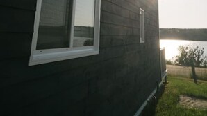 Une maison en bois sombre, avec deux fenêtres, donnant sur une étendue d’eau en arrière-plan.