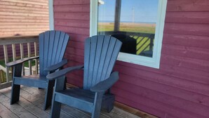 Two Adirondack chairs on a wooden deck with a view of a grassy field through a window. - Beach, Beach, Beach - Cabaneaux 1 (Chéticamp)