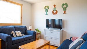 A cozy living room with a blue sofa, a wooden coffee table, a TV on a white cabinet, and decorative wall art. - Beach, Beach, Beach - Cabaneaux 1 (Chéticamp)
