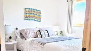A bedroom with a bed, a bedside table, a lamp, a wall hanging, and a window with curtains. - Beach, Beach, Beach - Cabaneaux 1 (Chéticamp)