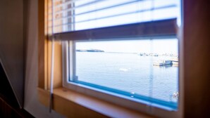 A window with horizontal blinds, a view of a calm body of water, and a distant shoreline with buildings. - The most wanted place to stay in Cheticamp (Chéticamp)