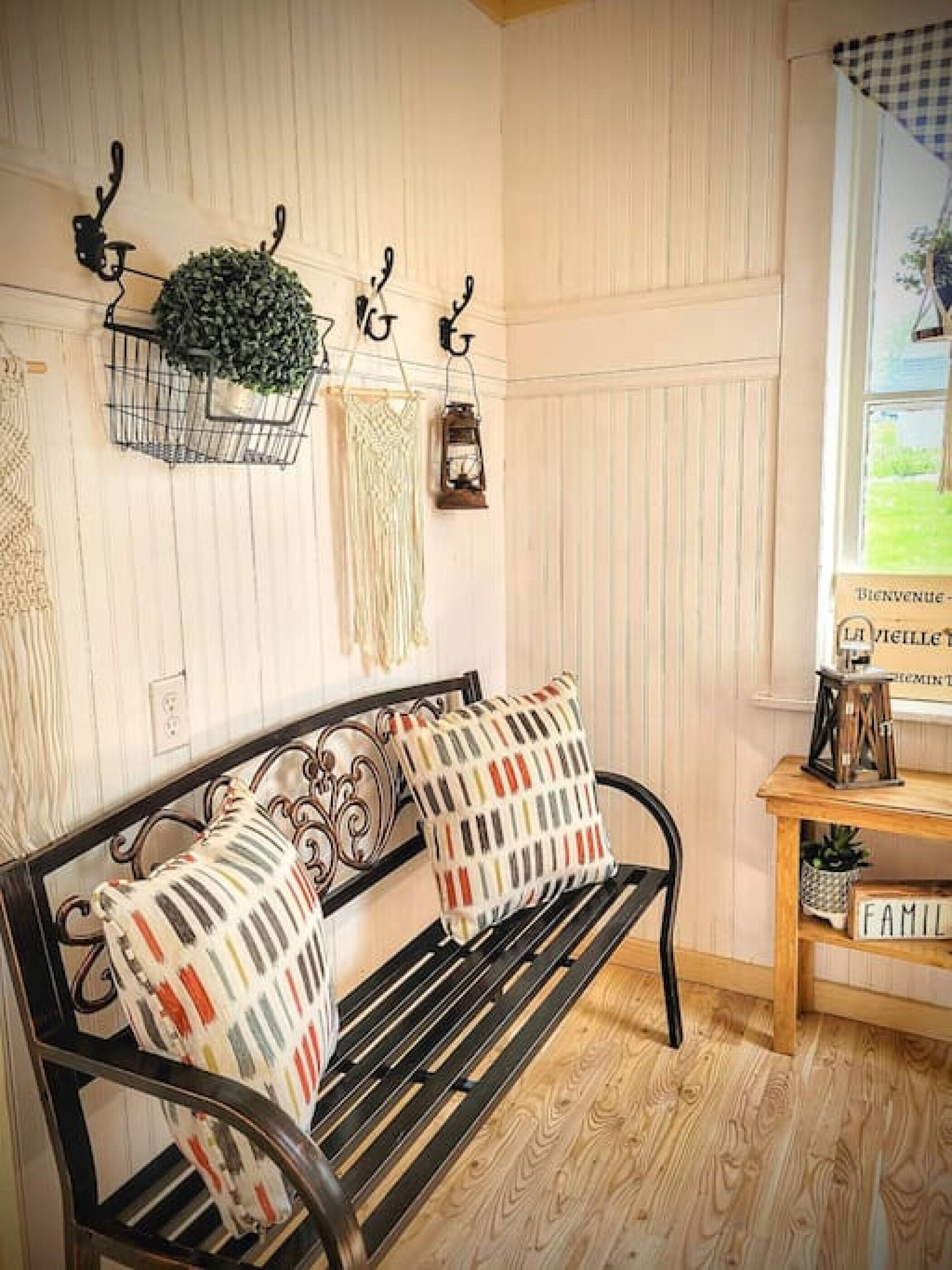 A wooden bench with patterned cushions, a wall-mounted plant, a lantern, and a welcome sign.
