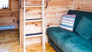 A wooden cabin interior with a loft bed, a green sofa, and a ladder to the upper bunk. - Tiny home on the Boardwalk (Chéticamp)