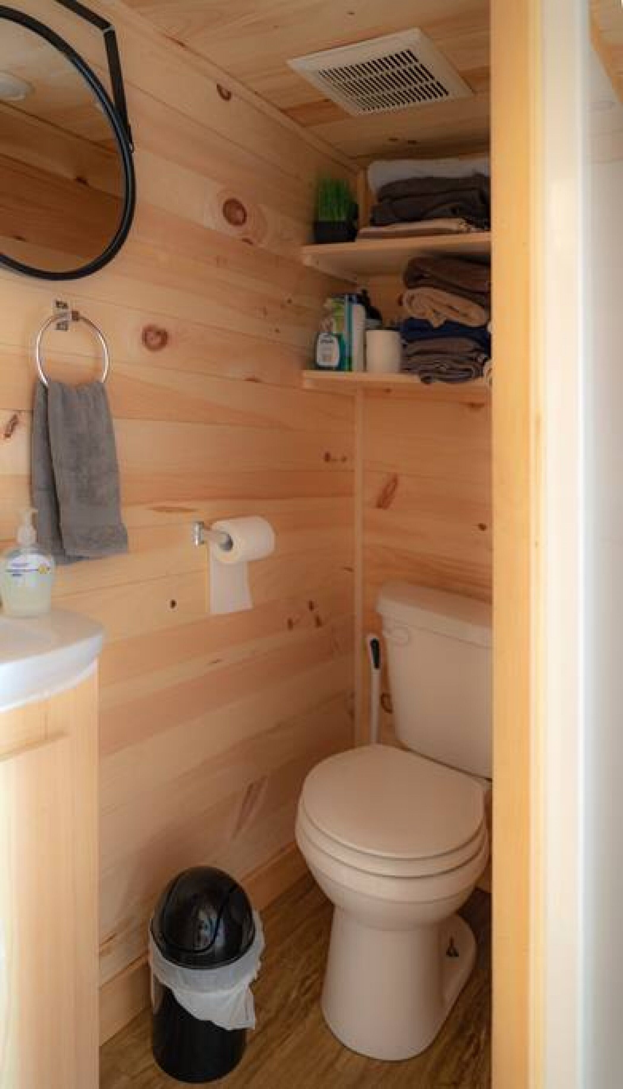 A compact wooden bathroom with a toilet, a sink, and a small shelf with toiletries.