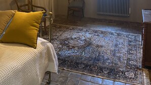 Una habitación con alfombra estampada, una cama con almohada amarilla, una silla de madera, una lámpara colgante y una ventana con vistas.