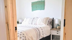 A bedroom with a bed, bedside tables, a lamp, and a green wall hanging. - Sunsets, barbecue`s and sandy beach - Cabaneaux 4 (Chéticamp)