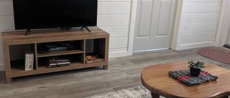 Een woonkamer met een flatscreen televisie op een houten standaard, een grijze bank, een ronde salontafel met een plant en een kamerkleed met een patroon.