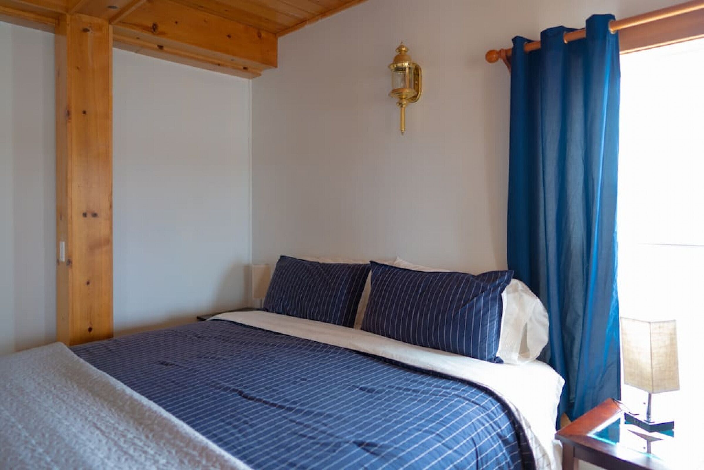 Une chambre à coucher avec un lit, des rideaux bleus, une lampe fixée au mur et un plafond en bois.