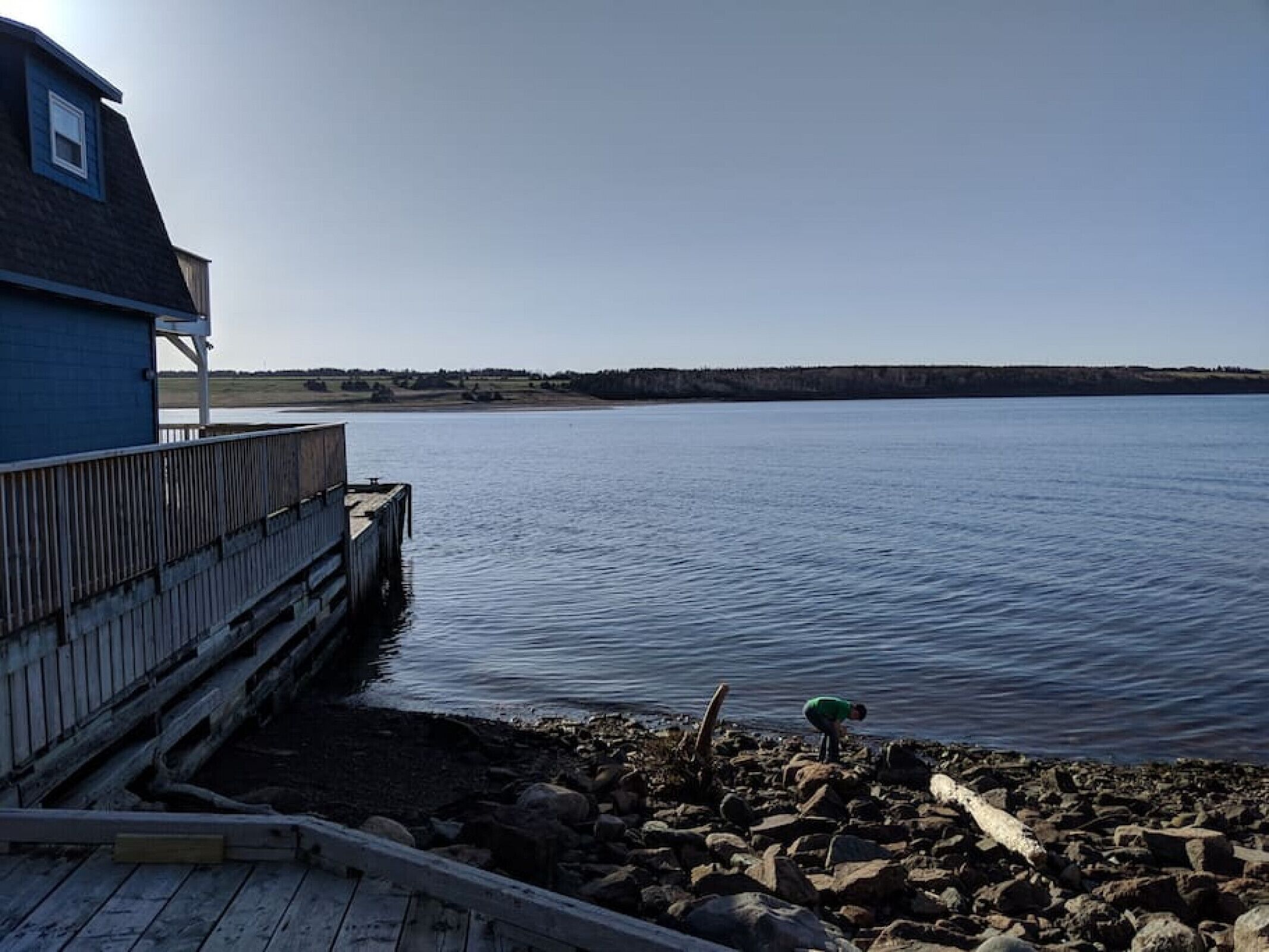 Eine Holzsteganlage, die sich in ruhiges Wasser erstreckt, ein Haus mit blauem Dach und eine felsige Küste.