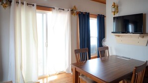 Uma  área de refeições com mesa e cadeiras de madeira, televisão fixada na parede e cortinas nas janelas.