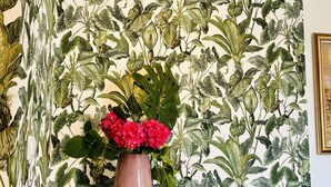 Una habitación con papel tapiz estampado, un armario de madera y un jarrón con flores rojas.