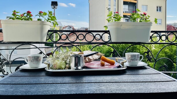 Un balcón con una mesa lista para el desayuno, plantas en macetas y vistas a edificios.