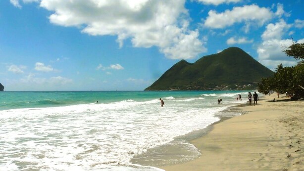 Une plage à l’eau turquoise et limpide, un rivage sablonneux et une montagne en arrière-plan.