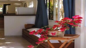 Une pièce avec un lit, une petite table sur laquelle est posé un vase de fleurs rouges, et une fenêtre avec des rideaux bleus.