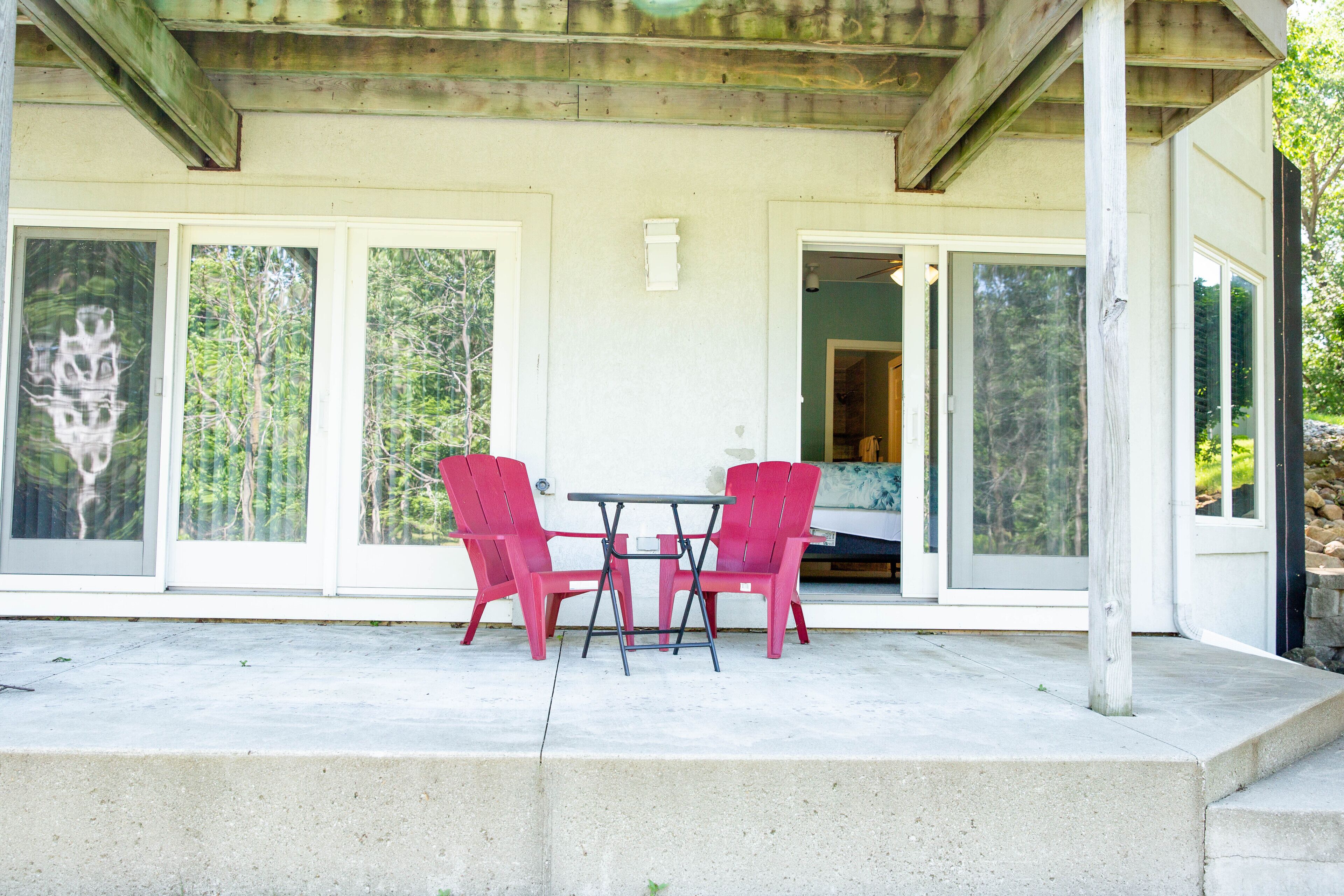 Un patio techado con una mesa de hormigón y dos sillas rojas, puertas de cristal grandes que dan a una habitación, y una estructura de vigas de madera.