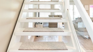 Een witte trap met een bekleed plateau, leidend naar een slaapkamer met een bed en een raam.