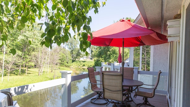 Een houten terras met een tafel en stoelen, een rode parasol en uitzicht op een waterpartij.