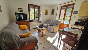 A living room with a sofa, armchair, coffee table, and a dining table with chairs. There is a television on a stand, a radiator, and a window with a view of greenery. - Le gite à mamie (ORBEY)