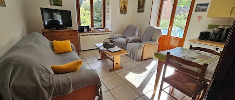 A living room with a sofa, armchair, coffee table, and a dining table with chairs. There is a television on a stand, a radiator, and a window with a view of greenery.