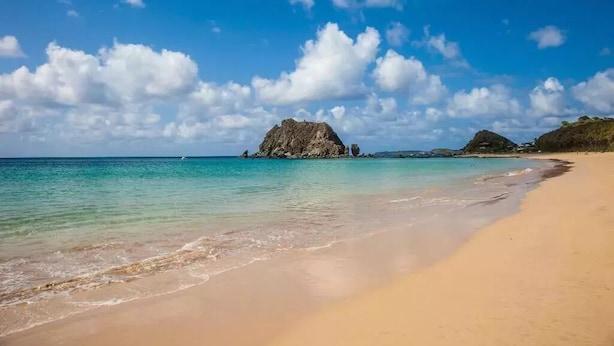 A beach with clear turquoise water, a sandy shore, and rocky formations in the distance under a blue sky with scattered clouds. - Pousada Maramar (Fernando de Noronha)